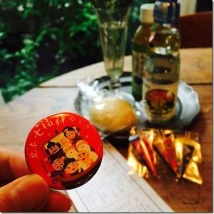 七福神の缶バッチを持った手と、テーブルの上に乗った飲み物と食べ物の写真