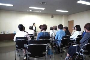 8名の会員が椅子に座り女性の話を聞いている写真