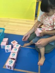 手作りの四角の箱を手に持ちお母さんの膝に抱っこされている女の子の写真