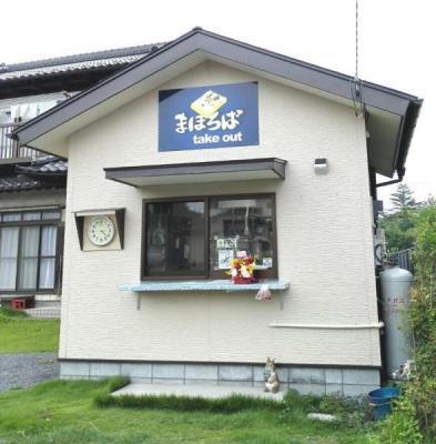 白い壁に屋根の下には黄色のひらがな文字でまほろばと書かれた青い看板と、受け渡しする為の出窓と横には丸い時計のあるまほろば外観写真