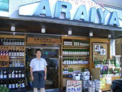 お酒が並ぶ店頭の前に立つ、腰にエプロンをまいたあらい屋の主人の写真