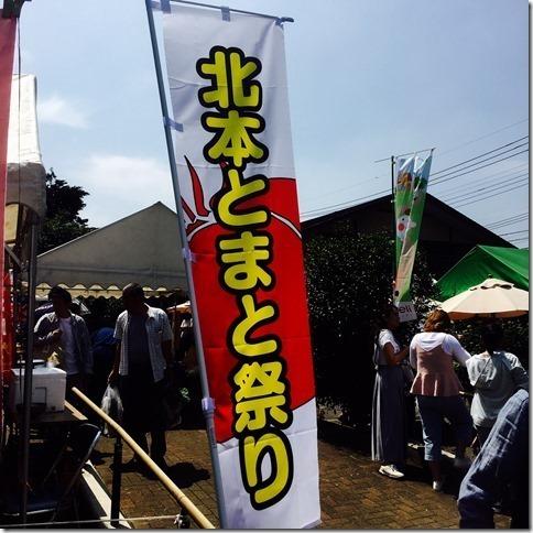 とまと祭りの旗と後ろのテントに人が集まっている写真