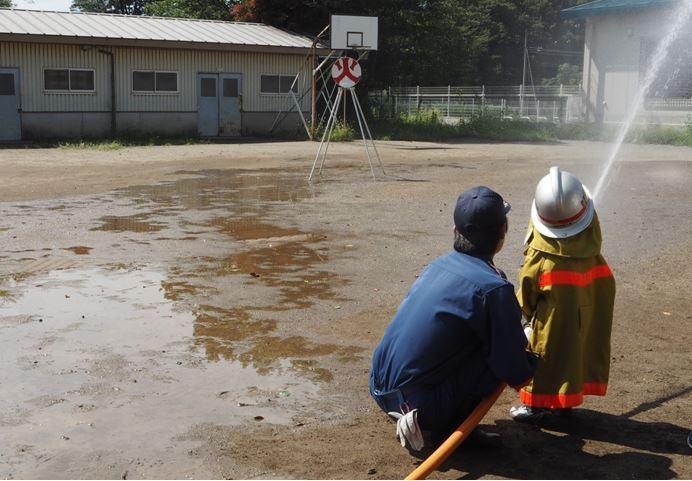 消防団員と一緒に、防護服とヘルメットをかぶって放水している子供の写真
