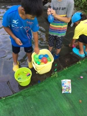 公園の水辺で男の子たちがバケツ一杯の水風船を作っている写真