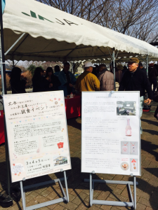 白いテントの下に集まるたくさんの人々と、手前には2枚の試食イベントを案内する看板の写真