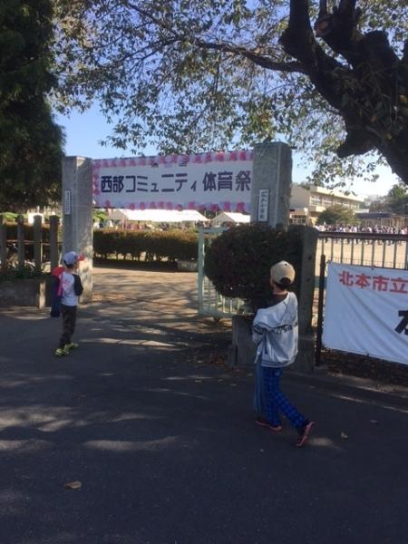 グラウンドの入り口に「西部コミュニティ体育祭」と大きく書かれた看板が設置されており、男の子2人が入り口に向かって歩いている写真