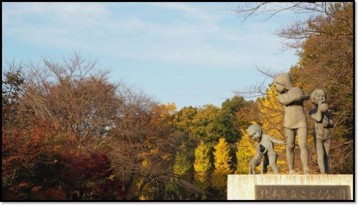 右手前に北本市立こども公園と書かれた台の上に3人の子供の像があり、奥に黄色や赤に色づいた紅葉の写真
