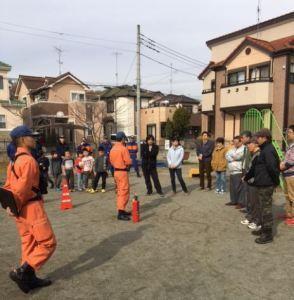 市民の皆さんが公園に集まっており、オレンジ色の消防服を着た隊員の話を聞いている写真