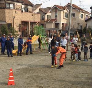 オレンジ色の消防服を着た隊員と小さな子供が一緒に放水訓練をしており、その様子を見ている市民の方々や青色の消防服を着た隊員の方々の写真