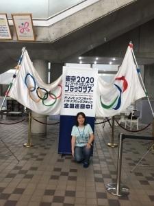 オリンピックの旗とパラリンピックの旗の間にあるフラッグツアーの看板の前で立膝をついている女性の写真