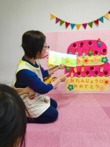 壁に貼ったケーキの前で絵本を読み聞かせている女性と、聞いている子供の写真