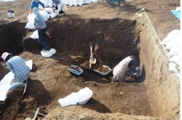 掘られた大きな穴の中で、作業員が、シャベルを使い土のう袋に詰めている写真