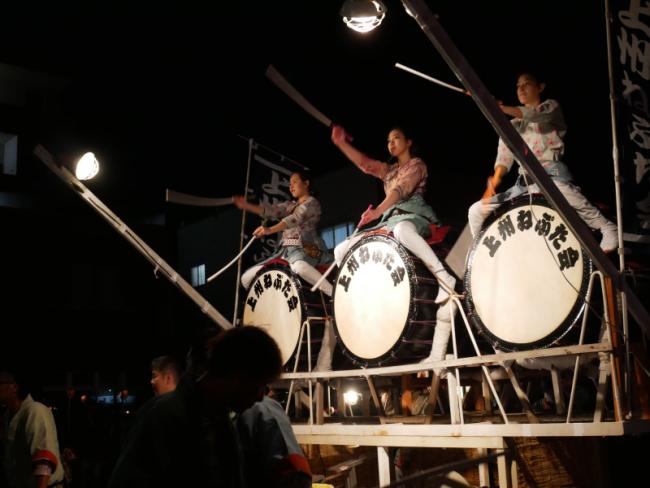 3人の女性が大きな丸い太鼓の上に乗って太鼓を叩いている写真