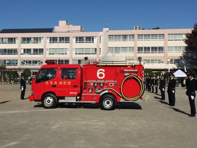 側面に6と書かれた消防車の後で制服を着た消防団員が等間隔に間をあけて並んでいる写真