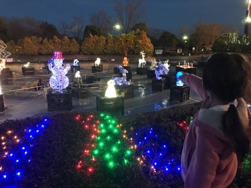 花壇越しに広場内に設置された雪だるまやトナカイのイルミネーションを女の子が指差している写真