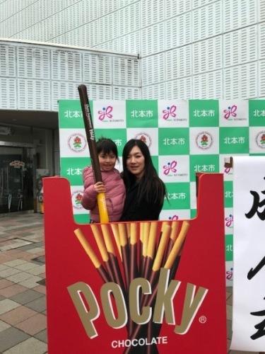 大きなポッキーの外箱の中で背丈くらいのポッキーを持った娘さんを抱っこするリポーターさんの写真