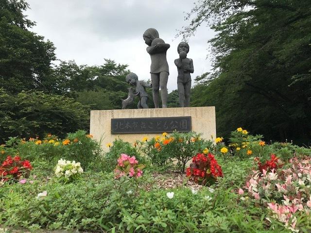 くもり空の下、中央にこども公園の3人の像と周りには色鮮やかな花々が咲き乱れている様子の写真