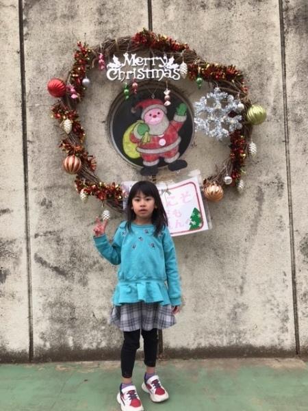 大きなクリスマスリースが飾られている前で水色の洋服を着た女の子がピースサインをしている写真