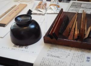 新聞紙の上に逆さまに置かれた漆塗りの黒いお椀と、漆塗りに必要なハケ等道具の写真