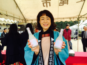 白いテントの下、両手に甘酒を持ってにこやかにほほ笑む北本市観光協会の水色の法被を着た山北さんの写真