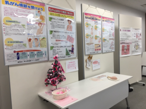 乳がん検査について記載されている展示物の前に置かれた机に、ピンクリボンの飾られたツリーが置かれている写真