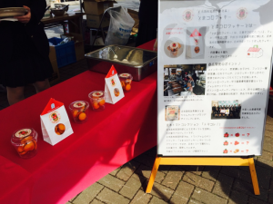 赤いテーブルクロスの敷かれた机の上に展示されているとまコロクッキーと、製作過程を説明した大きめの看板の写真