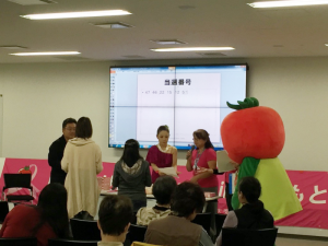 前方のモニターに当選番号が映し出されており、男性とピンク色の衣装を着た女性2人と着ぐるみを着たキャラクター「とまちゃん」が2名の女性に商品を手渡している写真