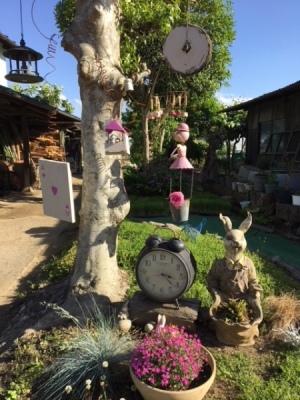 ピンクのお花とトランプや時計が吊るされている木とウサギの置物がある庭の写真