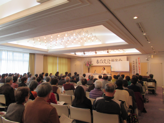 会場いっぱいに多くのお客様がいすに座り講演を聞いている写真