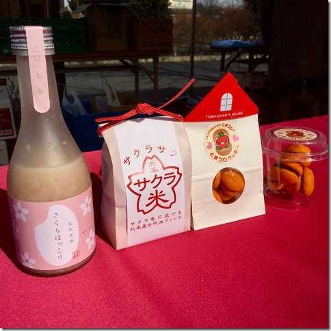 赤いテーブルの上に並べられた、手前からピンク色のラベルの甘酒、パッケージに桜が使われているサクラ米、白いパッケージのとまコロクッキーと透明なカップに入ったとまコロクッキーの写真