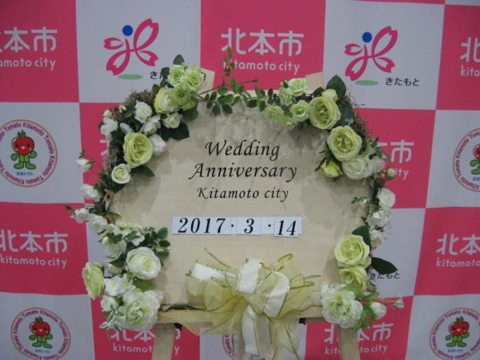 北本市のロゴが羅列されたパネルボードを背景とした、市が用意したウエディングボード（Wedding Anniversary Kitamoto Cityの文字と日付を記載し、周囲に白、グリーン系の花をあしらっています）の写真