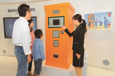 オレンジ色のグリコ自販機の案内をしている女性と、説明を聞いている親子3人の写真