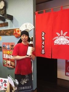 赤いTシャツを着た店主の佐藤さんが、赤ののれんの前で焼酎を持っている写真