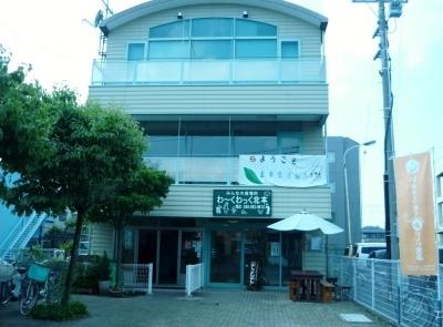外に白いパラソルと茶色のベンチが置いてあり、1階に大きな窓付きの入り口があるクリーム色の壁の3階建ての施設の写真