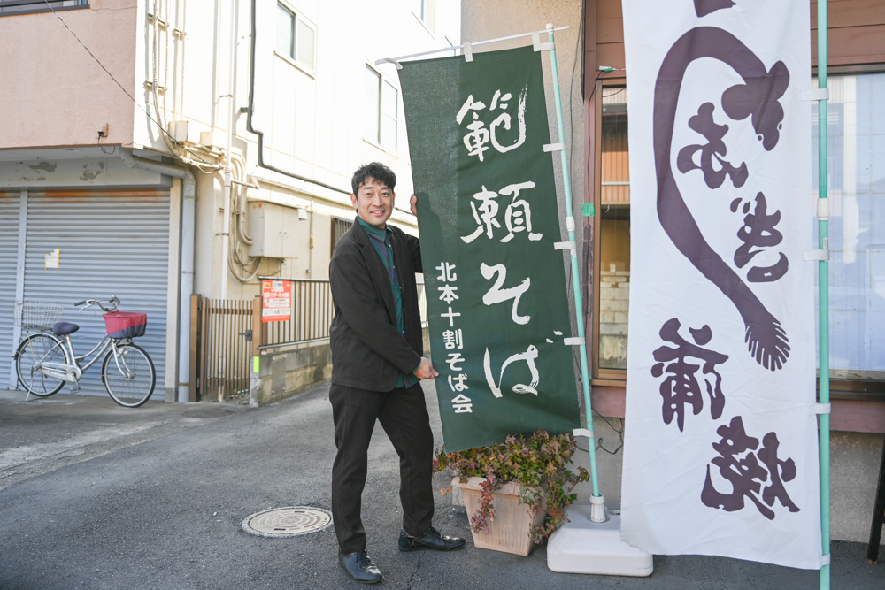 店の前で「範頼そば」と書かれたのぼりの横に立つ迫田さんの写真